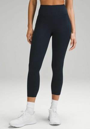 Leggings taille haute et ajustés en tissu bleu marine foncé. Ils présentent une texture lisse et une finition légèrement brillante, contrastant avec des chaussures de sport blanches.