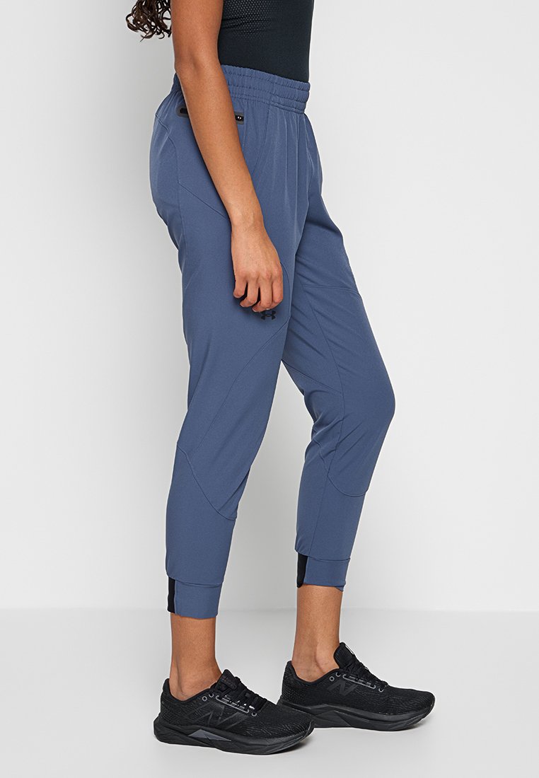 Femme portant un pantalon de jogging bleu et des chaussures de course noires, se tenant de côté contre un fond gris clair uni.