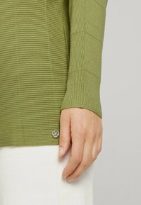 Grüner gerippter Strickpullover mit strukturiertem Design und einem kleinen silbernen Logoakzent am unteren Saum, kombiniert mit weißem Stoff.