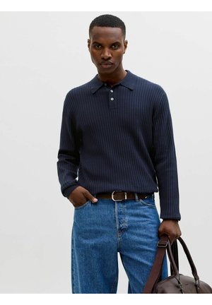 Homme portant un polo côtelé bleu marine à manches longues et un jean bleu, tenant un sac en cuir marron avec une sangle, debout devant un fond blanc uni.