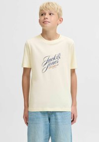 Plavi dječak s plavom kosom nosi kremastu Jack & Jones majicu i svijetloplave traperice, stoji i gleda lagano prema gore i udesno.