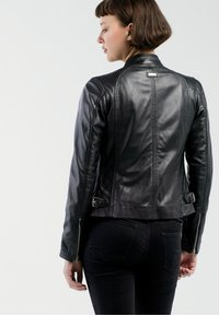 Schwarze Lederjacke, taillierter Schnitt, mit Reißverschlüssen an den Ärmelenden und zwei seitlichen Schnallen. Glatte Textur mit gesteppten Details auf dem Rücken.