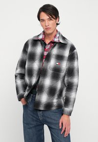 Vászonmintás, fekete-fehér pehelykabát, gombos elülső részével és logós mellzsebbel. Piros kockás inggel párosítva.