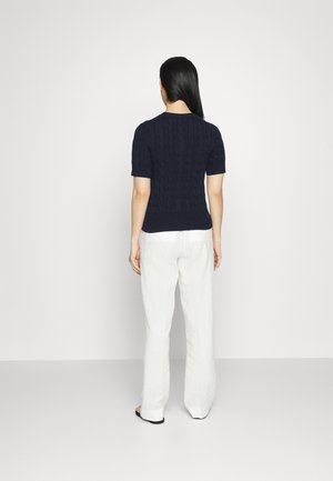 Femme aux longs cheveux foncés vue de dos, portant un pull en maille bleu marine à manches courtes et un pantalon blanc à jambes larges sur un fond uni.