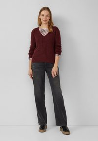 Frau, die einen bordeauxfarbenen Strickpullover über einem gestreiften Hemd trägt, schwarze weite Jeans und schwarze Stiefel mit braunen Sohlen, steht vor einer grauen Wand.
