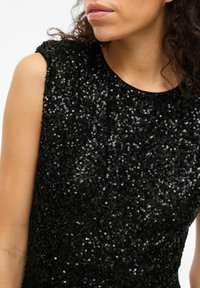 Woman wearing a sleeveless black top covered in small shiny sequins, with curly dark hair and lips visible, turned slightly right.