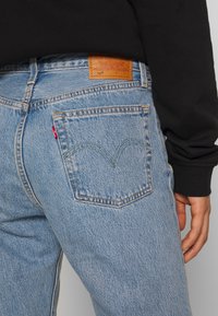Light blue denim jeans with a classic five-pocket design, featuring a brown leather tag on the back, and a stitched arcuate logo on the pocket.