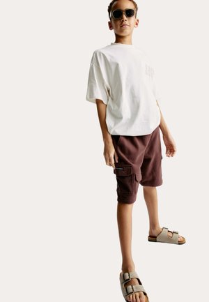Bílé oversized tričko, hnědé cargo šortky s bočními kapsami a béžové sandály s dvojitými přezkami, na neutrálním pozadí.