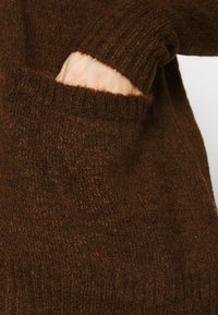 Maglione marrone lavorato a maglia con una finitura morbida e testurizzata. Presenta un colletto a coste e una tasca frontale posizionata sul lato inferiore.