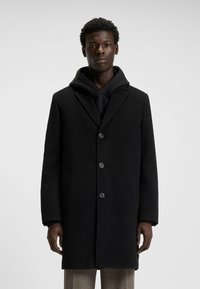 Cappotto di lana nero con colletto a punte, design monopetto e quattro bottoni. Sovrapposto a un hoodie nero.