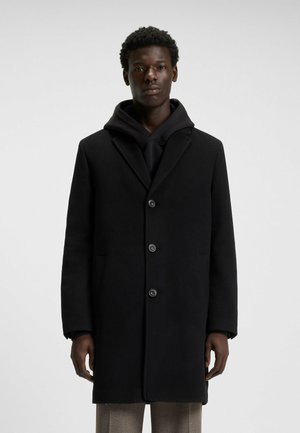 Black wool overcoat with a notched collar, single-breasted design, and four buttons. Layered over a black hoodie.