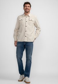 Helles beiges Cordhemd mit einer Knopfleiste, zwei Brusttaschen und dunklen Knöpfen, kombiniert mit blauen Jeans und grauen Turnschuhen.