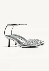 Silver spännande sandaler med en slank klack, öppen tå och ankelrem; har en strukturerad metallfinish och geometriska utskärningar.