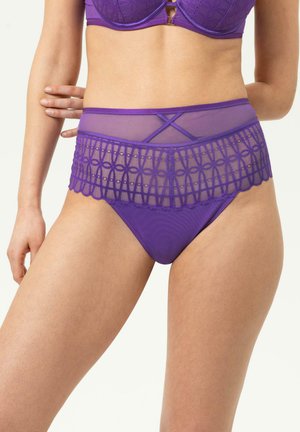 Vrouw draagt paarse kanten hoge taille ondergoed en bijpassende bh, hand rust opzij, tegen een effen lichte achtergrond.