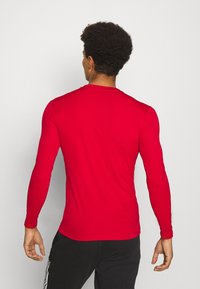 Langarmige rode sportshirt met een zachte, lichte textuur, ronde halslijn en een aangesloten ontwerp, gedragen met zwarte broek.