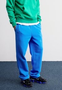 Hanorac verde cu buzunar tip cangur, asortat cu pantaloni de trening albaștri cu o textură netedă. Adidași negri cu accente albastre completează ținuta.