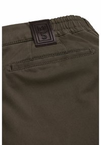 Khaki broek met een gestructureerd oppervlak, elastische tailleband en een rechthoekig bruin leren label met "M5." Inclusief een achterzak.