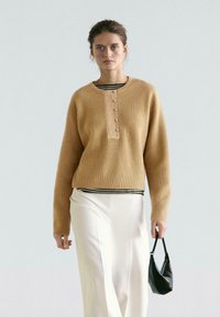 Pull côtelé beige avec un col rond, doté d'une patte de boutonnage. Porté sur un haut rayé et un pantalon blanc à jambes larges, accompagné d'un sac noir.