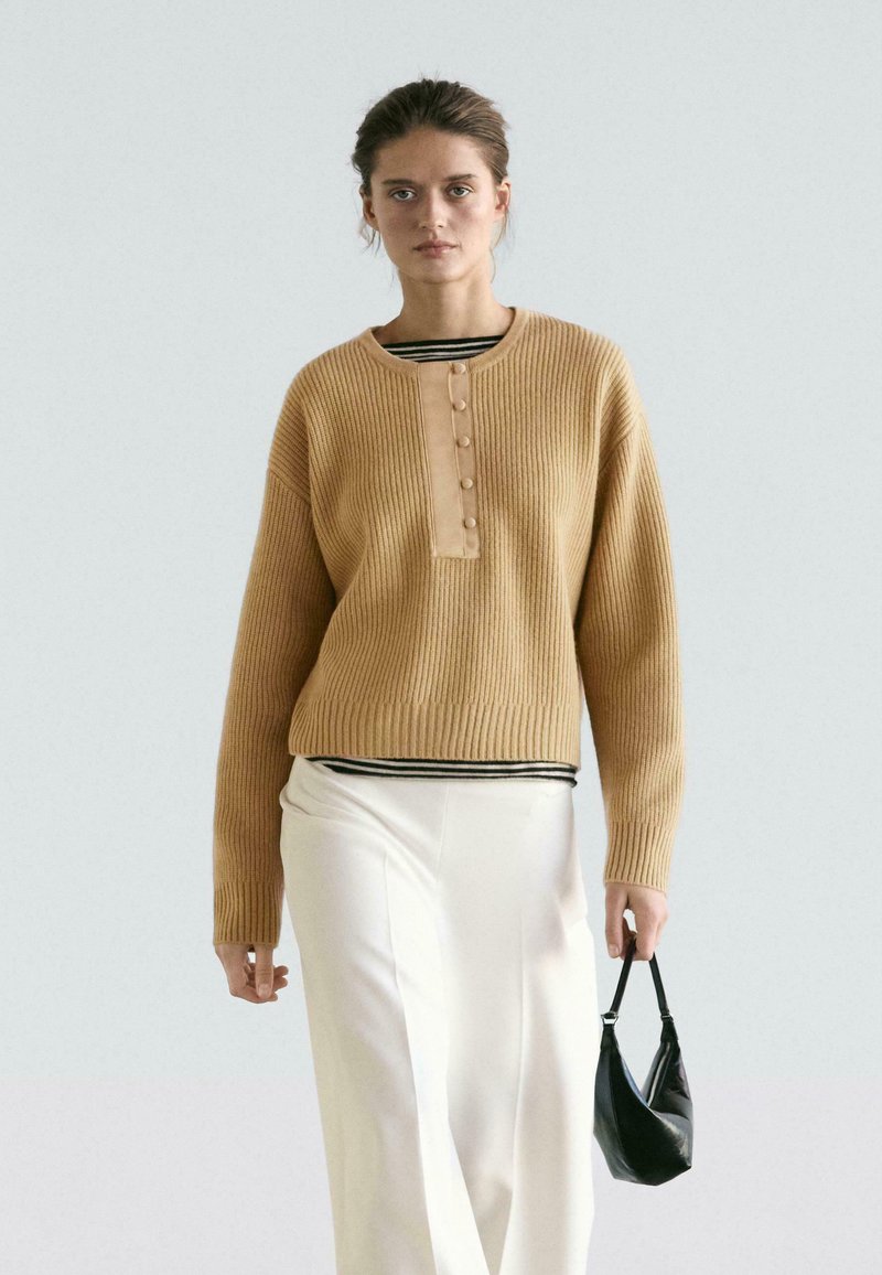 Pull côtelé beige avec un col rond, doté d'une patte de boutonnage. Porté sur un haut rayé et un pantalon blanc à jambes larges, accompagné d'un sac noir.