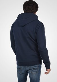 Navyblauwe hoodie met een trekkoordcapuchon, ribgebreide manchetten en onderrand. Glad stofoppervlak; model draagt het met blauwe jeans.