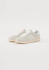 Hellgraue Wildledersneaker mit weißen Streifen und Schnürsenkeln, ausgestattet mit einer strukturierten Gummisohle und einem kleinen orangefarbenen Logo auf der Zunge.