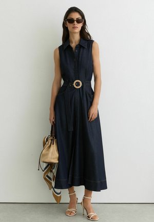 Kobieta w granatowej sukience bez rękawów z paskiem, beżowe sandały na wysokim obcasie, okulary przeciwsłoneczne, trzymająca beżową torebkę z apaszką na tle białej ściany.