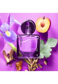 Fioletowa szklana butelka perfum z etykietą "KIKO MILANO COLONNE DISTRICT", otoczona płatkami irysa, plasterkiem jabłka i różnymi przyprawami na fioletowym tle.