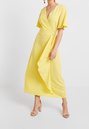 Woman wearing a yellow wrap dress with flutter sleeves and beige ankle-strap block heels, cropped above the neck against a plain background.