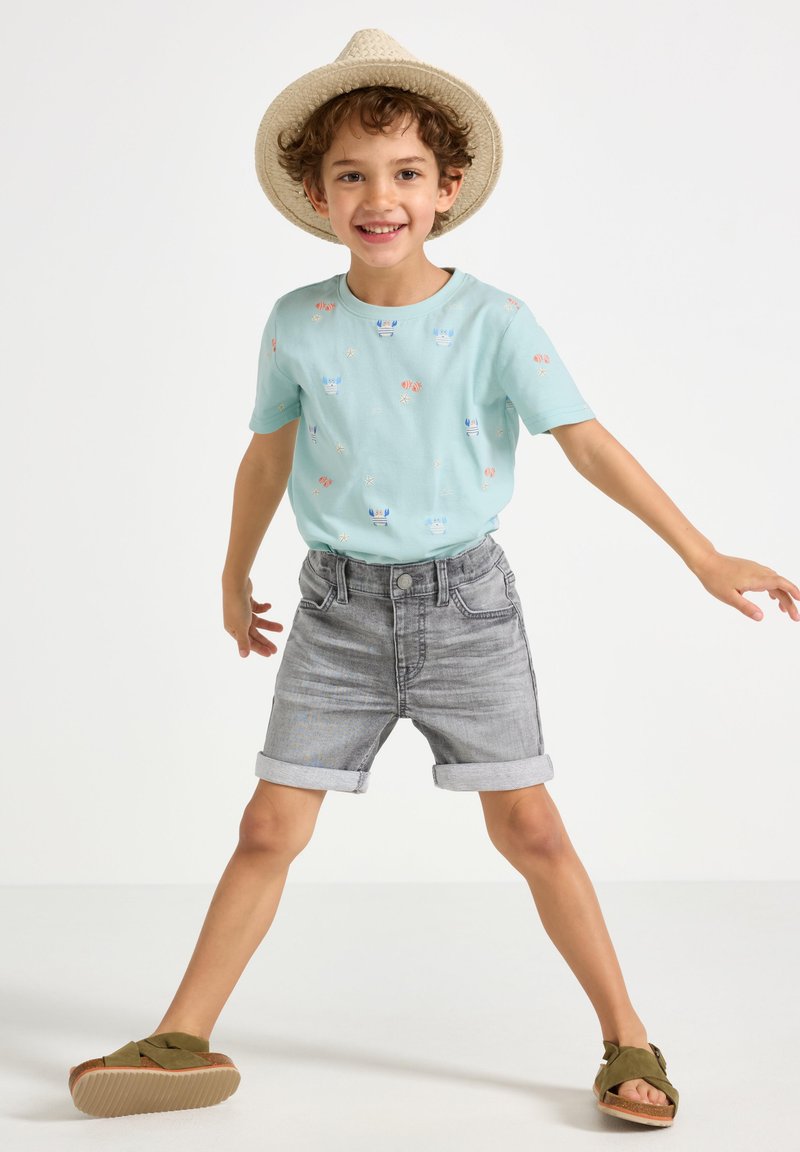 Lyseblå t-shirt med mønstrede prints, grå denim shorts med oprettede manchetter og grønne slip-on sko. Barnet poserer legende.