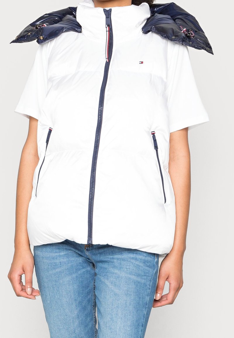 Biała kamizelka puchowa z wysokim kołnierzem, granatowymi akcentami na ramionach, z przodu na zamek błyskawiczny i bocznymi kieszeniami, zestawiona z białą bluzką z krótkim rękawem i niebieskimi dżinsami.