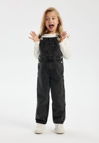 Black denim overalls with large front pocket, adjustable shoulder straps, and two side pockets, worn over a white long-sleeve shirt.