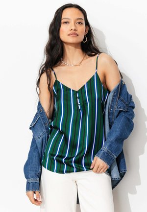 Femme aux longs cheveux foncés porte un caraco rayé verticalement vert, bleu et blanc, un pantalon blanc, avec une veste en jean bleue tombant sur les épaules.