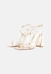 Πέδιλα με ψηλό τακούνι σε μεταλλικό χρυσό φινίρισμα, με μαργαριταρένιες λεπτομέρειες και διαφανή σχεδιασμό λουριών. Τετράγωνη μύτη και χοντρά τακούνια.