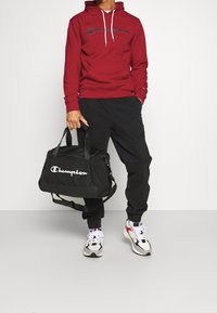 Sweat-shirt à capuche rouge avec le logo "Champion", sac de sport noir avec logo blanc, pantalon de jogging noir, et baskets multicolores avec détails blancs.
