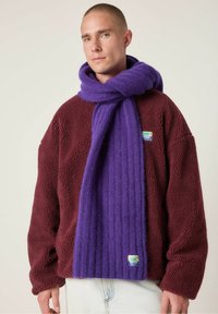 Vijoličasti rebrasti šal iz mehkega materiala, nosi se z maroon puhastim puloverjem. Oznaka vidna na puloverju z podatki o blagovni znamki. Enostaven in teksturiran dizajn.