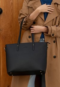Zwarte leren tote bag met dubbele handvatten, gestructureerde vorm, textuurafwerking en metallic hardware accenten, die rust op een houten achtergrond.