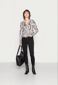 Blusa de mangas compridas com um padrão ondulado em bege, castanho e azul, combinada com jeans skinny pretos e botins, segurando uma bolsa preta.
