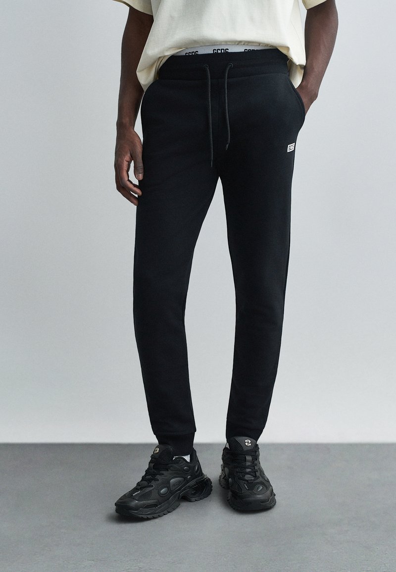 Black sweatpants with elastic waistband and drawstring, featuring a logo patch on the hip. Paired with black sneakers on a gray floor.