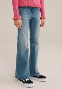 Rozšířené džíny z denimu v světle modré barvě, s hladkým povrchem a mírně vybledlým vzhledem. Kombinované s černými teniskami s bílými tkaničkami.