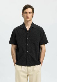 Chemise noire à manches courtes avec un motif texturé en tourbillon, dotée d'un col et de boutons sur le devant ; le modèle porte un pantalon crème.