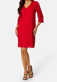Robes en dentelle rouge avec un décolleté en V, des manches trois-quarts et un ourlet festonné. Présente des motifs floraux et une coupe ample. Associée à des sandales noires à lanières.