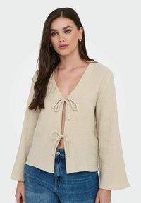 Beige Bluse mit vornem Bindeband, langen Ärmeln, strukturiertem Stoff und tiefem V-Ausschnitt. Getragen über blauen Jeans mit legerem Schnitt.
