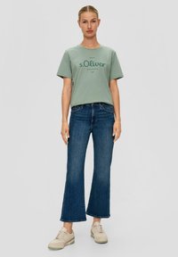 Groene katoenen t-shirt met donkerdruk, gecombineerd met blauwe flared jeans en lichte sneakers. Eenvoudig, casual ontwerp met korte mouwen.
