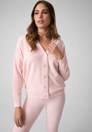 Femme portant un cardigan rose pâle doux boutonné et des leggings assortis, touchant le col du cardigan, sur un fond gris clair uni.