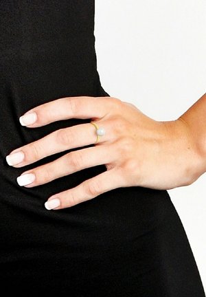 Bague en or avec une perle blanche lisse sur le dessus, portée sur une main avec une manucure française, sur fond de tissu noir.