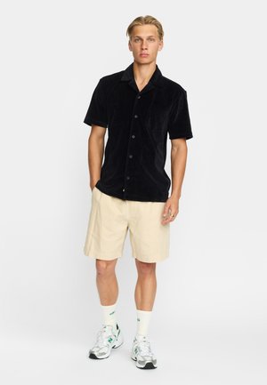 Schwarzes kurzärmliges Hemd aus weichem Stoff mit einer Tasche, kombiniert mit hellbeigen Shorts und weißen Turnschuhen mit grünen Akzenten.