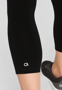 Czarne legginsy o dopasowanym kroju, krótkiej długości i z małym białym logo na dolnej stronie. Gładki, elastyczny materiał.