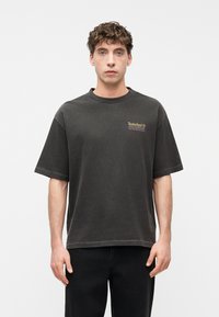 Νεαρός με σγουρά μαλλιά που φοράει μια κοντομάνικη μπλούζα Timberland σε σκούρο γκρι χρώμα και μαύρο παντελόνι, στέκεται μπροστά σε ένα απλό λευκό φόντο.