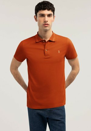 Mann trägt rostorangefarbenes Polohemd mit zwei Knöpfen, steht mit den Händen hinter dem Rücken vor schlichtem hellem Hintergrund.