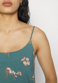 Top sin mangas de color verde azulado con patrones de flores y mariposas en tonos rosa y marrón, con una tela suave y un escote redondeado.